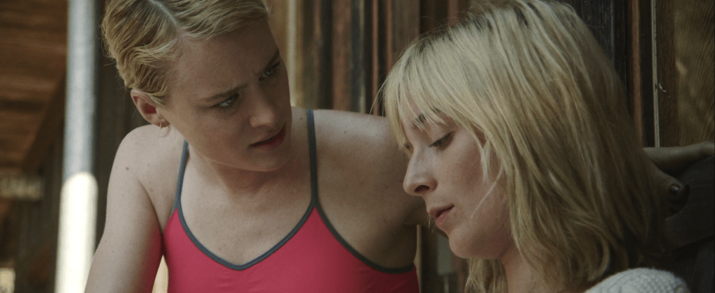 Image is a close-up of Anna, to the left, and Beth, to the right. Beth is looking down at her script that’s off-screen, her eyes not meeting Anna as if she doesn’t dare to. Anna, wearing a bright pink sports bra with a grey lining around it, is almost hanging over Beth. Anna is in full control, with a strained face looking right at Beth with her intense and piercing eyes as they’re running lines.  