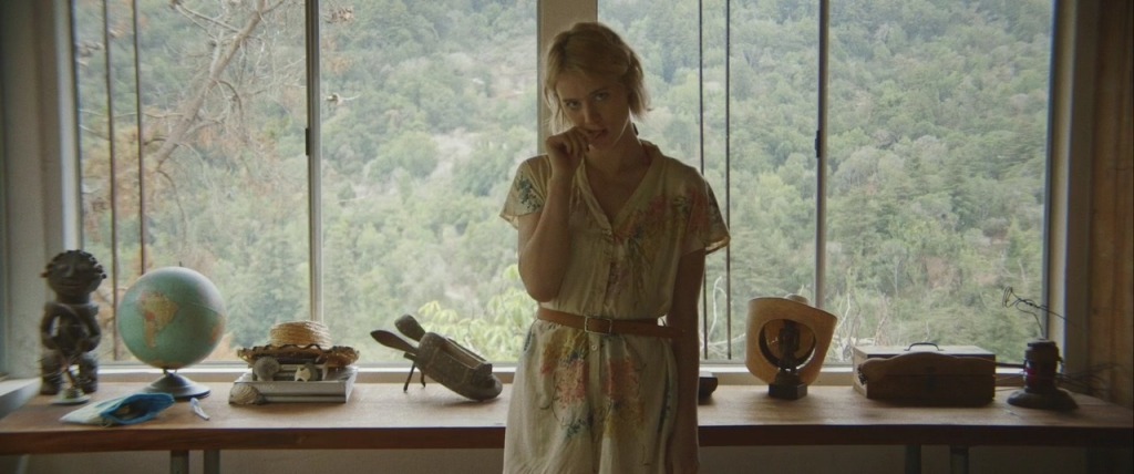 Image is from the film 'Always Shine' (2016). The film still shows Anna, now performing as Beth, in the middle of the frame. She’s looking right into the camera with her head tilted to one side, almost like she knows viewers are watching her performance. She’s biting on her thumb, reminiscent of an innocent child as she’s trying to mimic the body language of Beth. She looks nervous and timid. Anna is wearing Beth’s button-up romper suit with a flower pattern, with a light brown belt in the waist. In the background is a long desk visible – filled with miscellaneous objects including a globe and a cowboy hat all in a row – along with a big window showing the green nature of Big Sur.