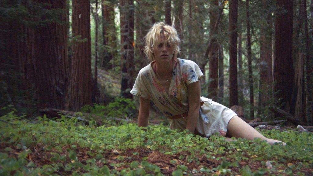 Image shows Anna – still looking like Beth in her physical appearance – waking up in the woods of Big Sur. She’s alone and she looks disoriented in her facial expression, with a confused expression as she looks ahead of herself with a mixture of emptiness and fear in her eyes. She’s still wearing Beth’s button-up romper suit with a flower pattern with a light brown belt in the waist.  