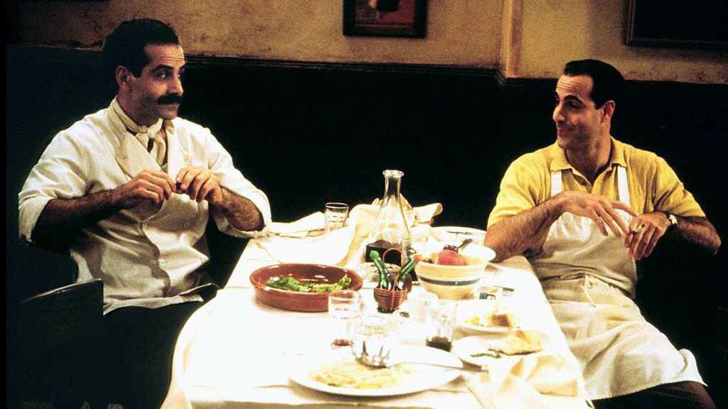 Image is from the frilm 'Big Night' (1996). Two chefs sit opposite one another with their hands to their chests and talk to one another. They sit either side of a white, clothed, table which is covered with half-eaten food.