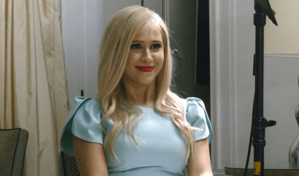 Image is from the film 'Borat Subsequent Moviefilm' (2020). A young woman wearing bright red lipstick and a pale blue dress smiles. 
