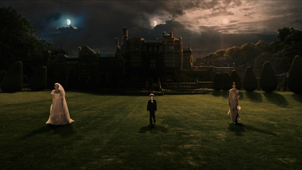 Three people stand in front of a shadowed castle that stands in front of a cloudy night sky that shows three moons. On the left is a woman wearing a wedding dress. In the middle is a child wearing a suit. On the right is a woman wearing a purple dress.