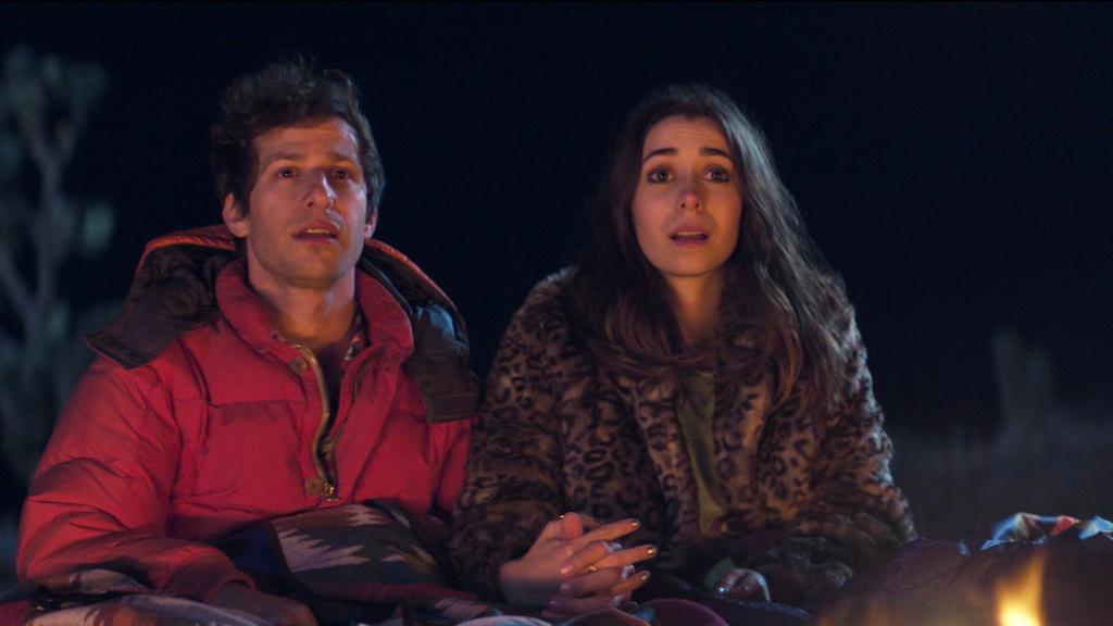Image is from the film 'Palm Spring' (2020). A man in a red coat holds the hand on a woman wearing a leopard print fur coat. They are sat in front of a fire at night, covered in blankets. They both are looking at something in the distance.