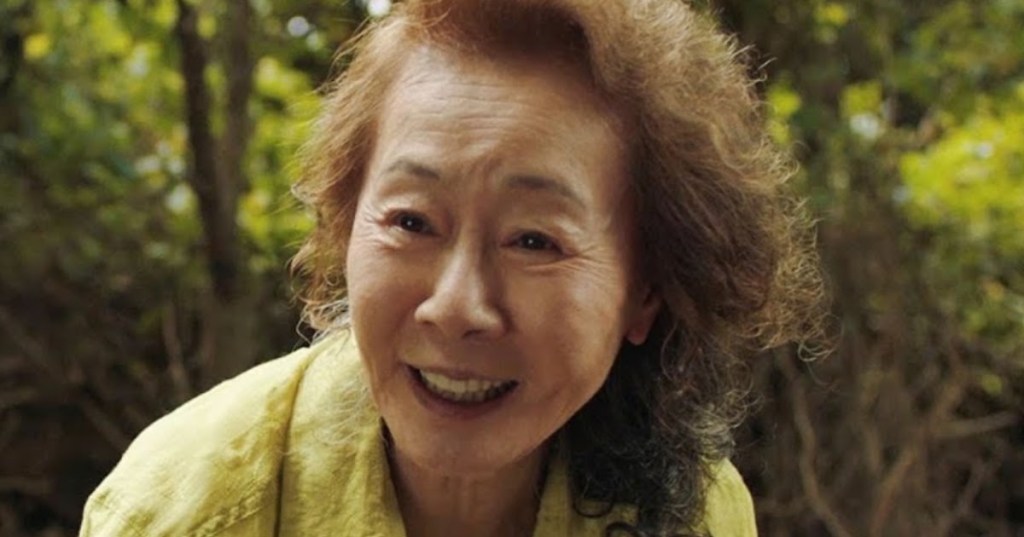 Image is from the film 'Minari'. An elderly woman in a forest leans down and smiles. She is wearing a yellow shirt.