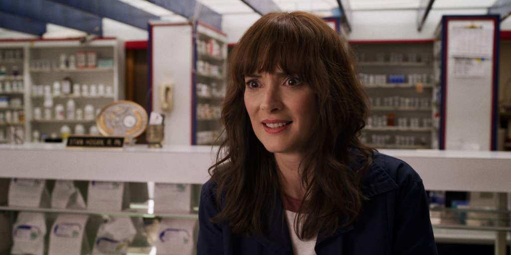 Joyce wears a concerned smiles in the middle of a pharmacy. She is wearing a blue coat and a white top.