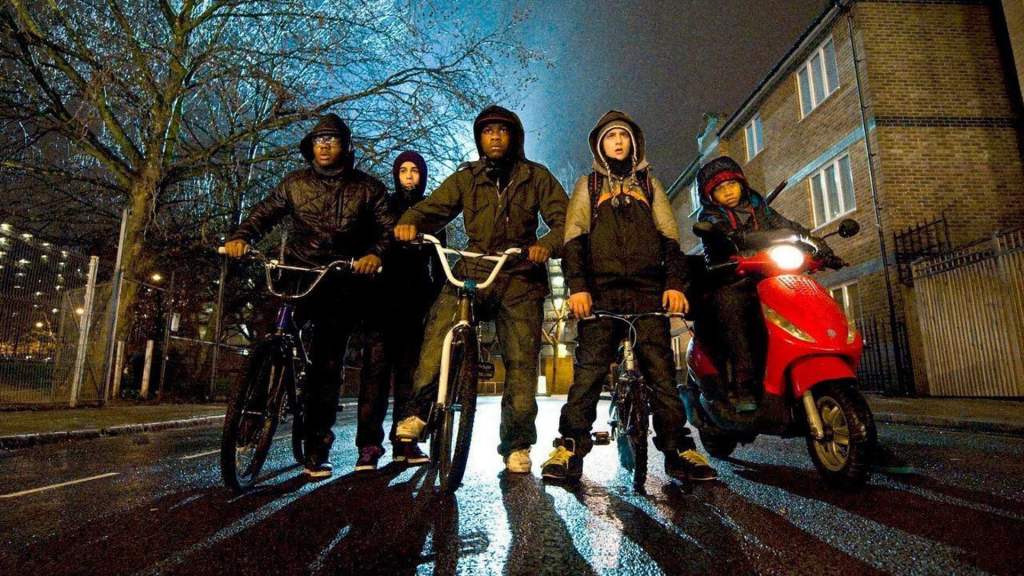 Image is from 'Attack on the Block' (2011). A group of teenagers on various bikes stand in the middle of a dark road and look shocked at something in front of them. Behind them, a blue light glows.