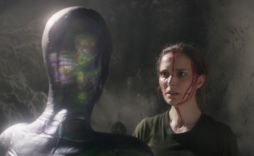 A woman wearing a dark green t-shirt with streaks of blood down her face and neck. She stares at a mysterious humanoid creature who has bright colours on the side of its head.