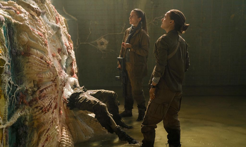 Two women dressed in dark green outfits look upon an alien life form. A pair of legs wearing matching pants that are ripped stick out from the bottom.