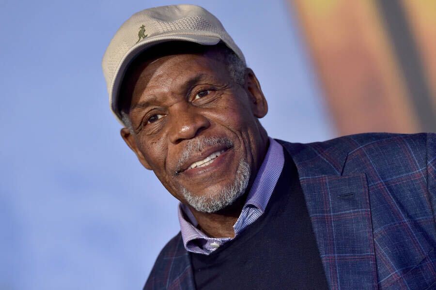 Danny Glover, a Black man in his 70s, smiles for the camera. He wears a navy plaid jacket, collared shirt, and tan baseball cap with a kangaroo.