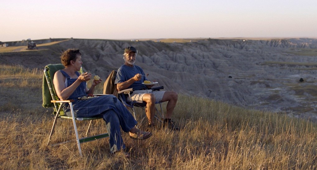 On the edge of a cliff, an older man and woman sit on deckchairs and eat a meal together. The landscape around them is highlighted in shadows and golden sun.