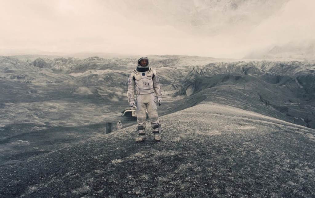 An astronaut stands in the center of a desolate landscape, with craters and an ashen grey sky.
