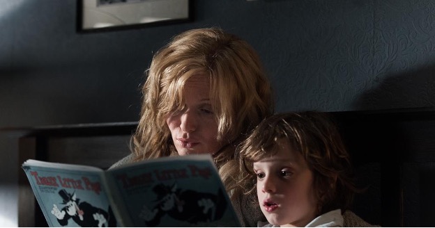 An older woman with blonde hair and a child sit together reading. The blue book cover in front of them has an illustration of a cartoon wolf in a fancy outfit and says 'Three Little Pigs'.