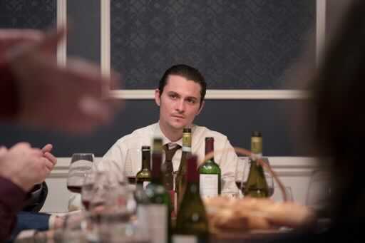 A man wearing a white collared shirt sits at a table. Bottles of wine are in front of him, and people's hands appear in frame.