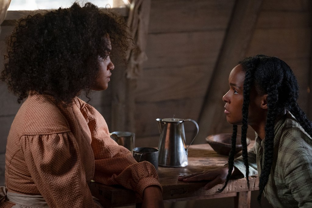 Two Black women sitting at a table and looking at one another. The woman on the left has an afro and wears a long-sleeved top, while the woman on the right has braids and wears a plaid shirt. A metal pitcher is seen on the wooden table between them.