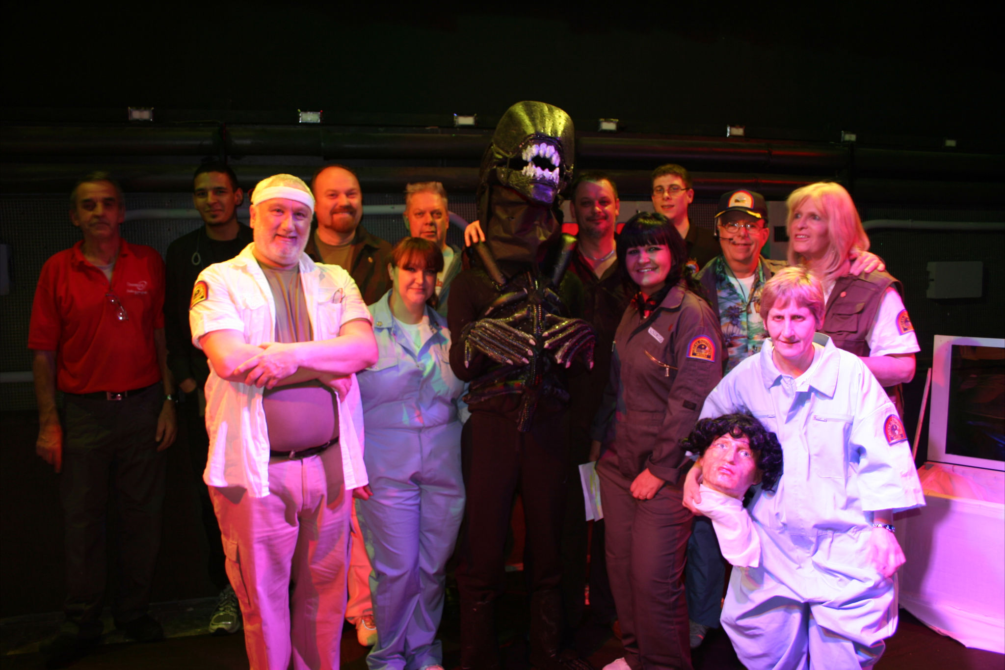 A group of middle-aged people, many of whom are dressed in costume as characters from the film 'Alien'. The actor playing the Alien stands in the centre, surrounded by others in spacesuits. A woman on the far right holds a severed head prop.