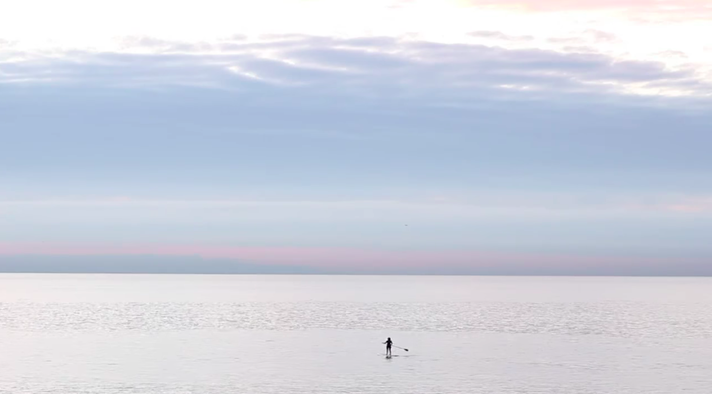 Marie (Jane Adams) way out on the ocean, standing on her board and paddling away from the camera alone. It's a wide static shot, perfectly framing the meeting of the ocean and sky horizontally. It feels never-ending. Colours of pink, orange, light and darker blue.