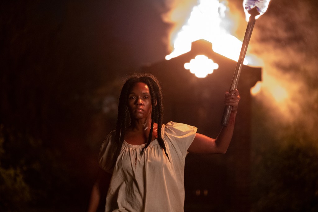 With a sullen look on her face, Veronica holds a burning torch walking away from a shed behind her that is lit on fire.