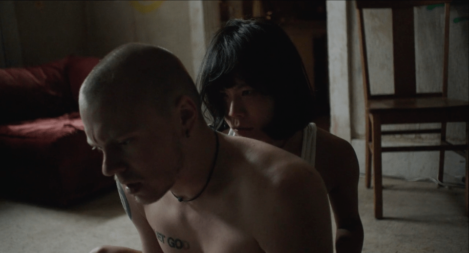 An Asian woman with short brunette hair sits behind a young man with a shaved head. He has a partial tattoo showing that reads "let go," and they are in a dingy apartment.