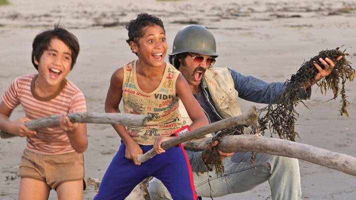 Two young boys wearing colourful summer clothes, with big smiles and holding branches. A man stands beside them, wearing a hat and an excited expression while holding up moss. All three of them are in front of a body of water.