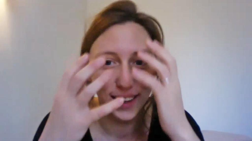 A young woman with brunette hair, as seen through a computer screen. She is smiling and her hands are in front of her face.