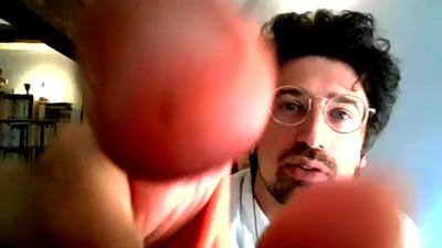 A man with dark hair, a moustache and beard, and glasses shown through a computer screen. His hand is in front of the camera in extreme close-up as he examines something.