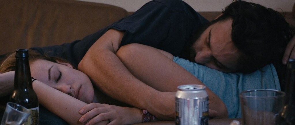 beer cans, bottles and glasses in the foreground but taking up the whole frame is Kate (Olivia Wilde) and Luke (Jake Johnson) who has fallen asleep intertwined with Kate on her side resting her head on Luke's lap and Luke resting his head on Kate's waist while holding his arm around her. It looks like they're captured an early morning after having one too many beers the night before. It's intimate, but in a way deeper than solely romance, as they're fully comfortable and safe with each other.