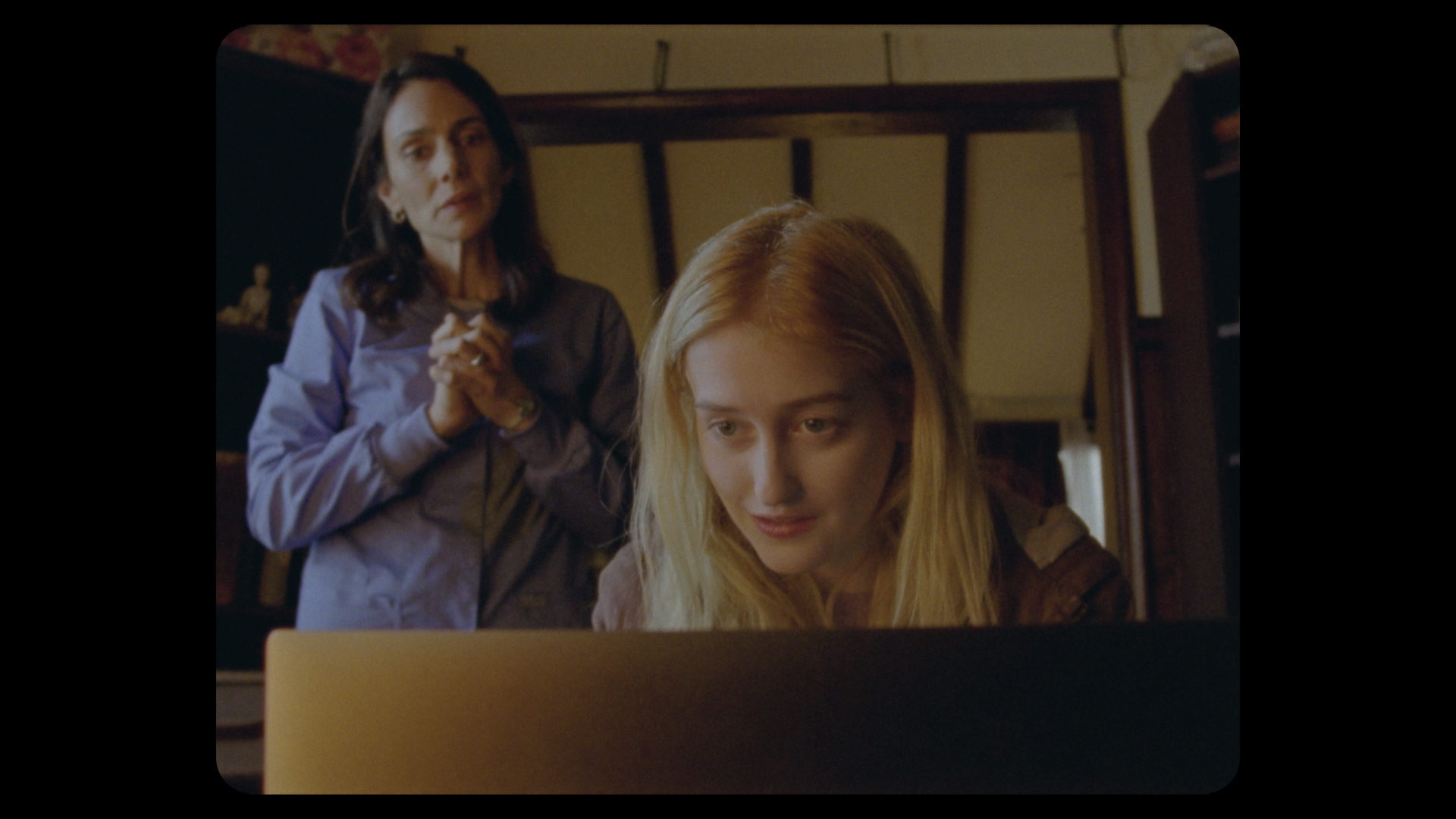 A young blonde woman sits behind a computer, smiling. An older brunette woman stands behind her, wearing a blue shirt. They are in an office space.