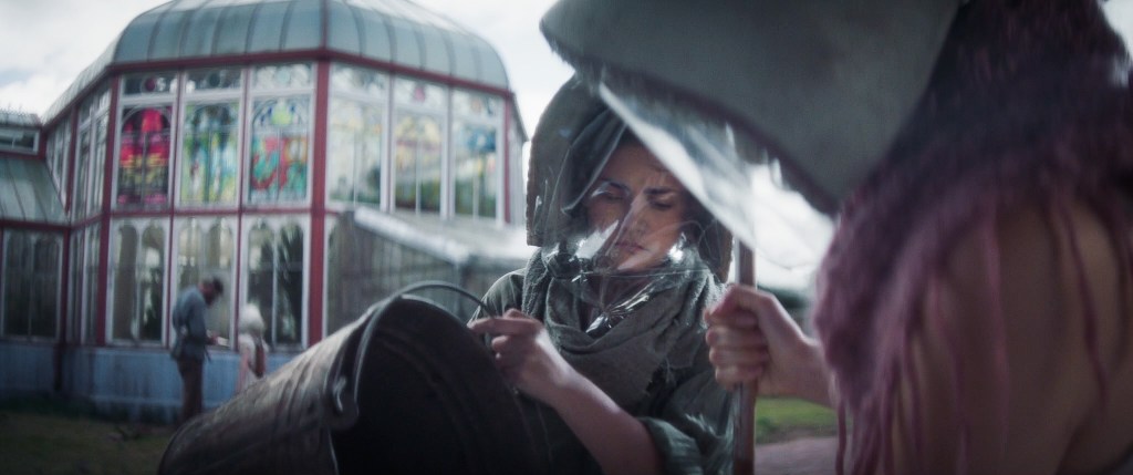 A young woman wearing a protective facial mask and loose clothing while holding a pail. A greenhouse with stained glass windows is out of focus behind her. Another person stands to her right wearing the same protective gear.