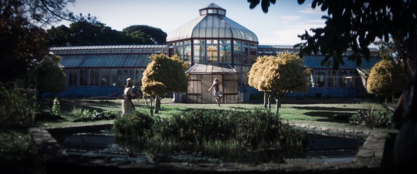 A greenhouse with stained glass windows visible from afar. Two figures can be seen from afar— one running in front of the building and the other outside. It is daytime, and they are surrounded by green foliage.