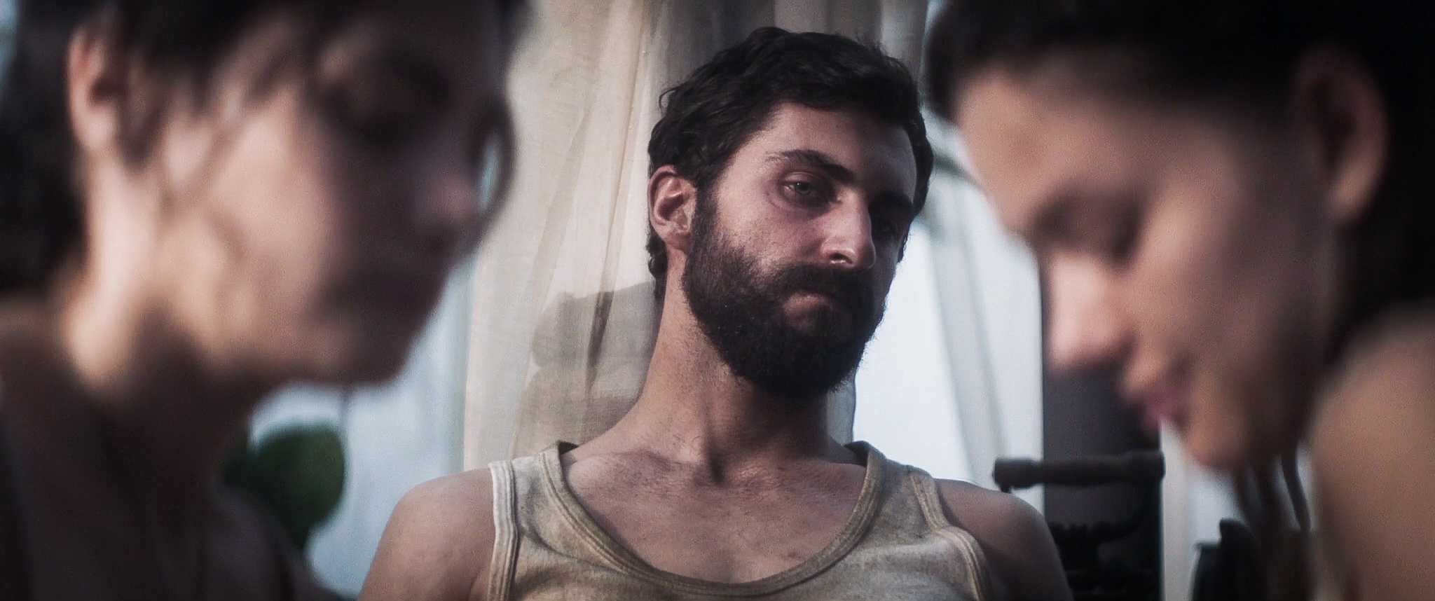 A bearded man with dark hair and a dirty tank top. He is visible from the chest up and between two young women in out of focus close up.