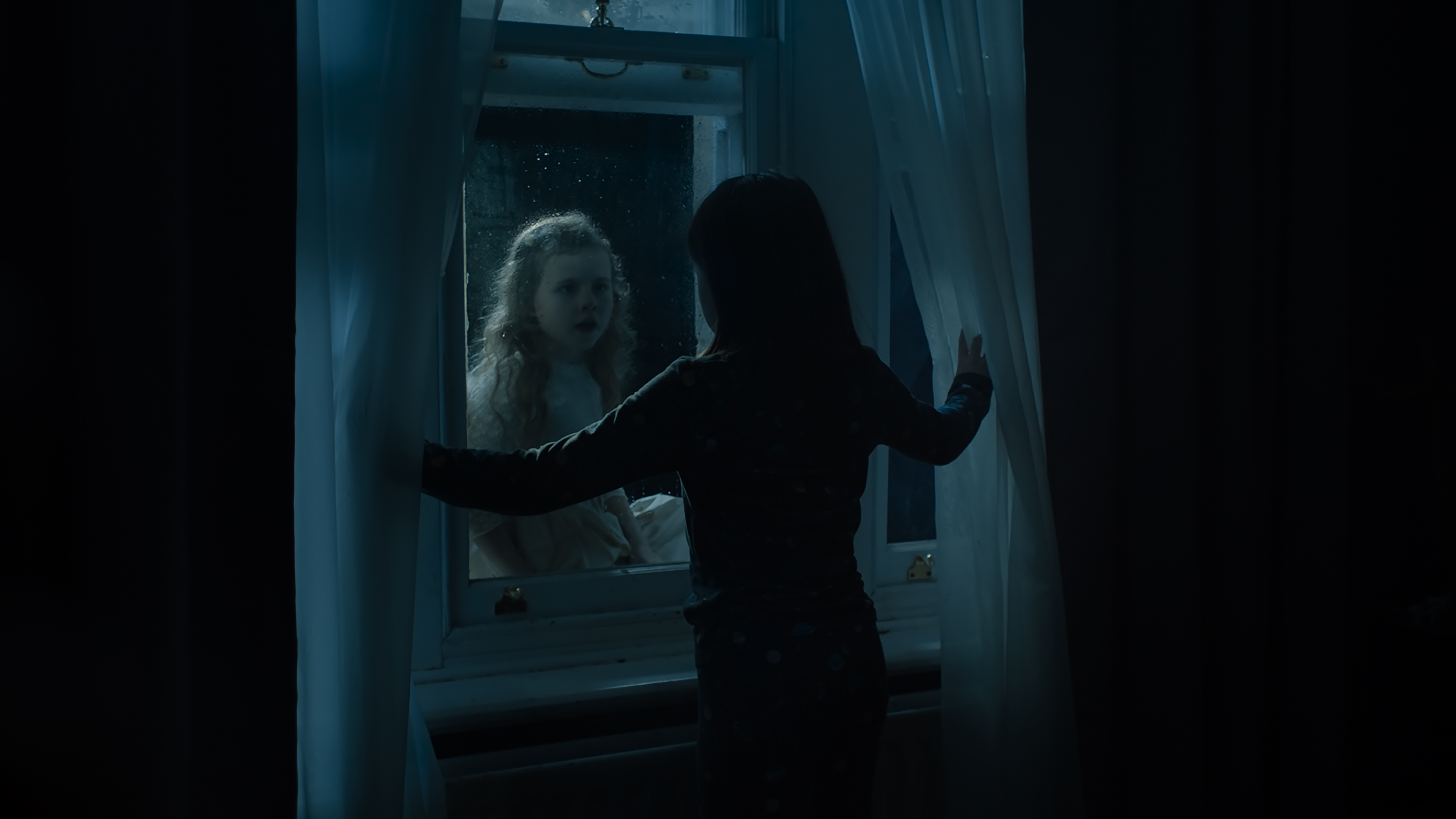 A young girl seen from behind in shadow standing in front of a closed window. A young girl with light hair and a white dress can be seen on the other side of the glass.