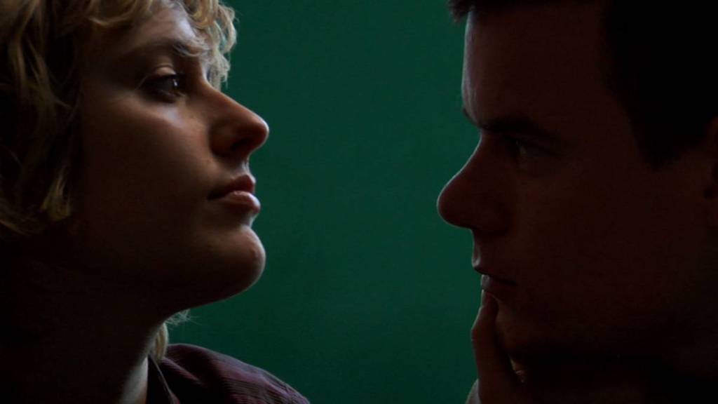 Close-up of James (Swanberg) and Mattie (Gerwig), Mattie to the frame's left and James to the right. It's a dark shot, with shadows darkening James' face, while light hits the highpoints of Mattie's face lighting up her chin, lips, cheekbones and nose. Mattie's head is lightly tilted upwards, in a stern position, as if she's the one in control in the frame. While looking at him, she holds James' face in her hand. James, with a more concerned facial expression, looks into her eyes.