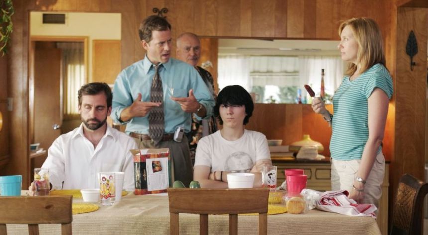 Two men in white shirts sitting at a kitchen table that has been set. The older man has a beard and neutral expression, while the younger man has dark emo-style hair and crossed arms. A man in a blue shirt stands between them angrily gesturing to a woman, who is wearing a blue striped shirt. An older man is also visible behind them.