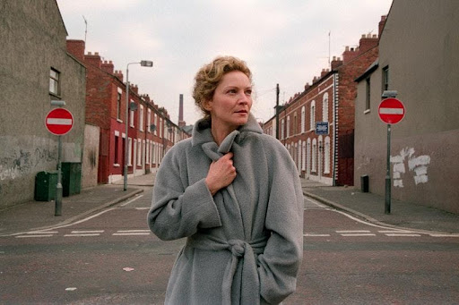 Courtesy of: Sally Potter/Sony Pictures Alt text: A woman stands centre frame, with a work-class street stretching out behind her. She is clutching her grey woollen coat around her and her face is unflinching.