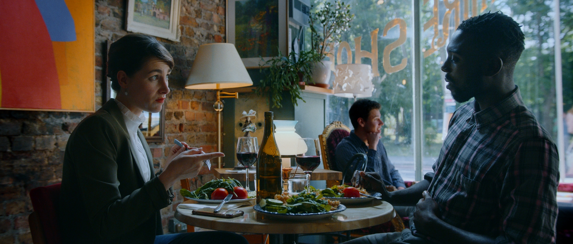 A young brunette Caucasian woman sits across the table at a restaurant from a Black man. She appears unimpressed, while his mouth is open. There is a table of food in front of them, as well as a brick wall with art and a window behind them with part of the writing . A man sits near the window.