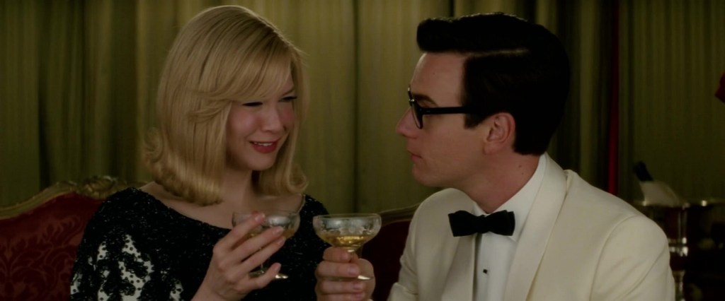 A man and a woman having drinks while sitting on a couch. They look at each other with drinks in their hands. The woman wears a dress and smiles, while the man wears a white tuxedo and has an unreadable expression.