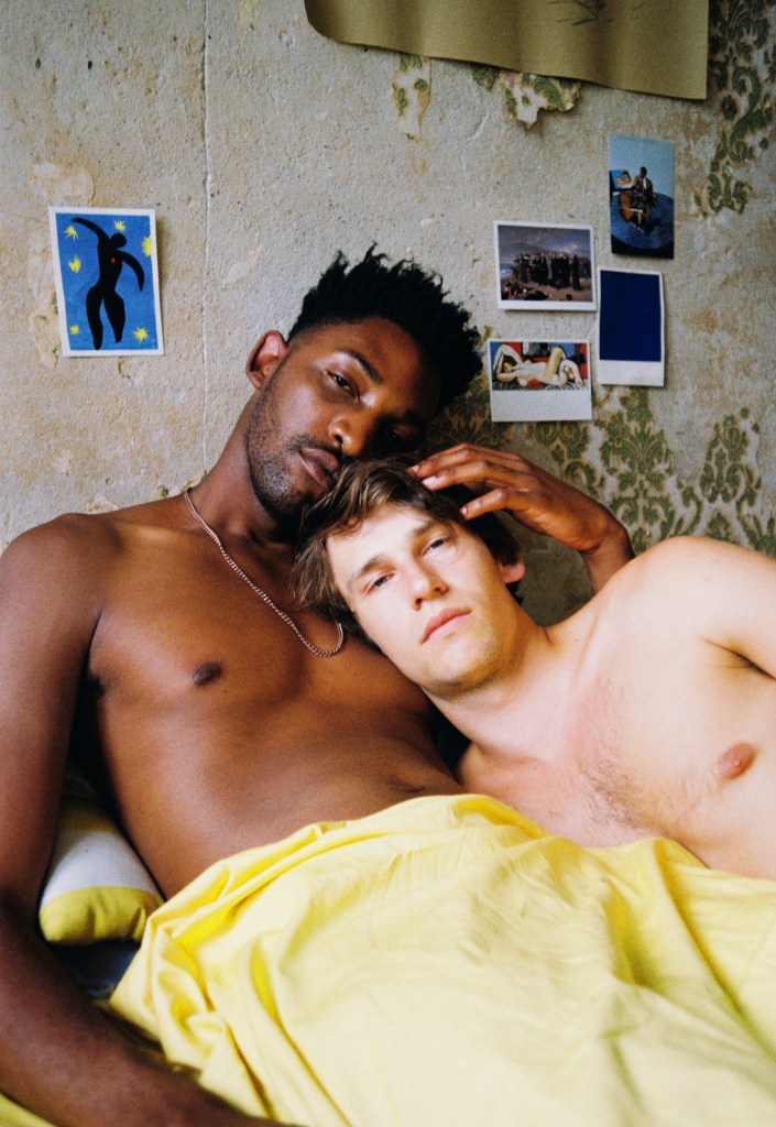 Harry and Johannes sit up in bed, covered by a yellow sheet. Harry holds Johannes' head, resting his head on top.