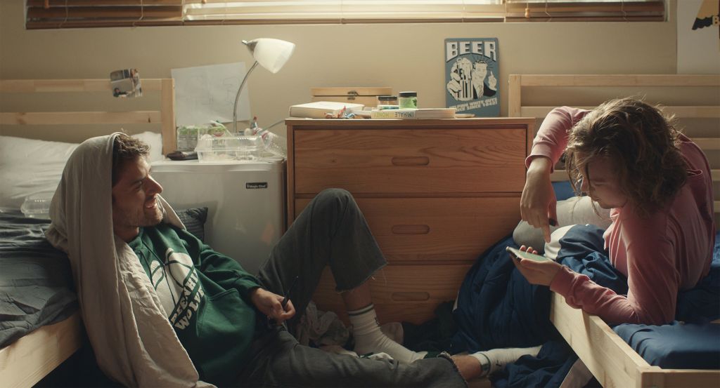 Two young men in a college dorm room. One has short brunette hair and wears a green hoodie with another hoodie draped over his head, leaning against a bed. The other has messy brunette hair and facial hair, wearing a light pink long sleeve shirt. He looks down, pointing to something on his phone.