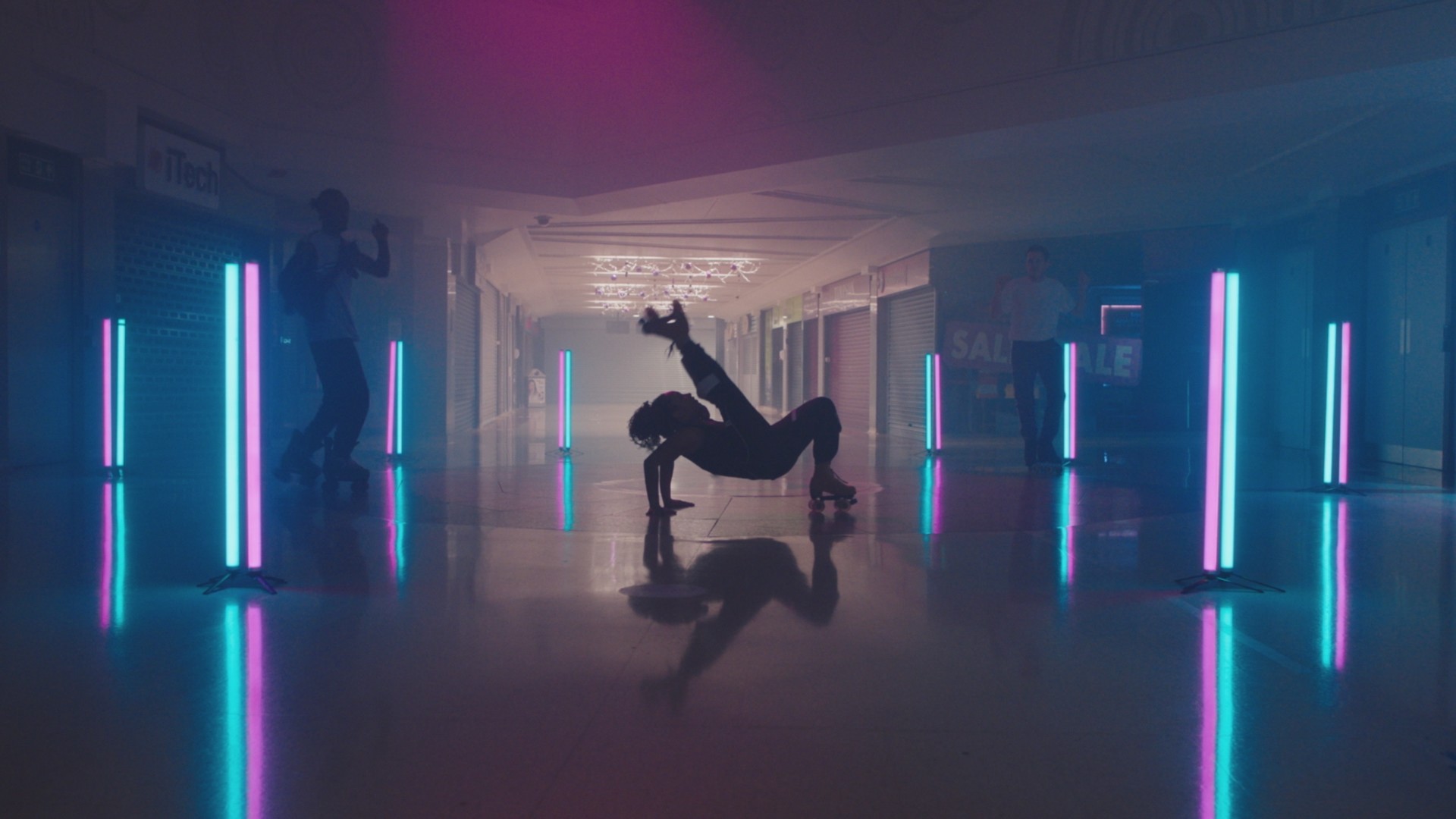 Young people on rollerskates in an empty mall. The shot is illuminated by neon pink and blue lighting.