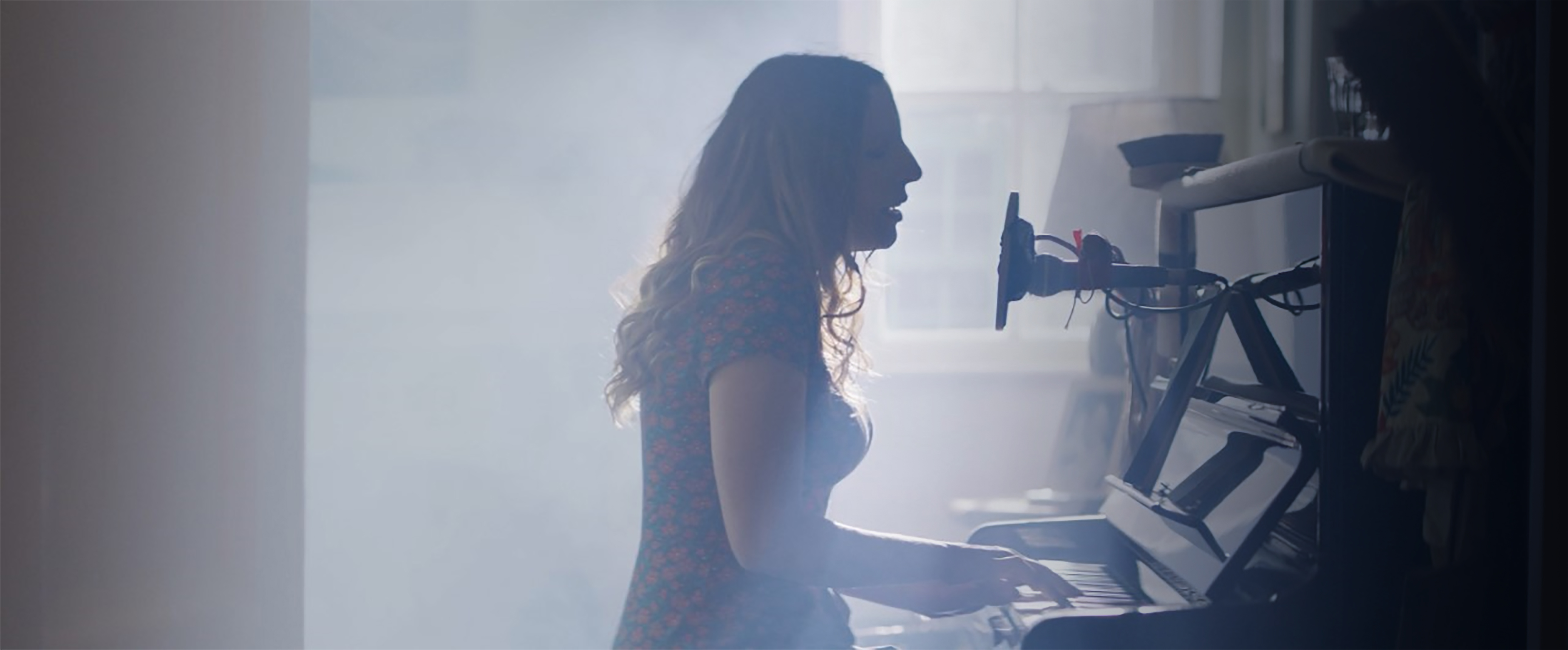 A silhouette of a dark-haired woman singing and playing the piano. She is seen from the side, and has a makeshift recording setup, including a creatively fashioned mic.