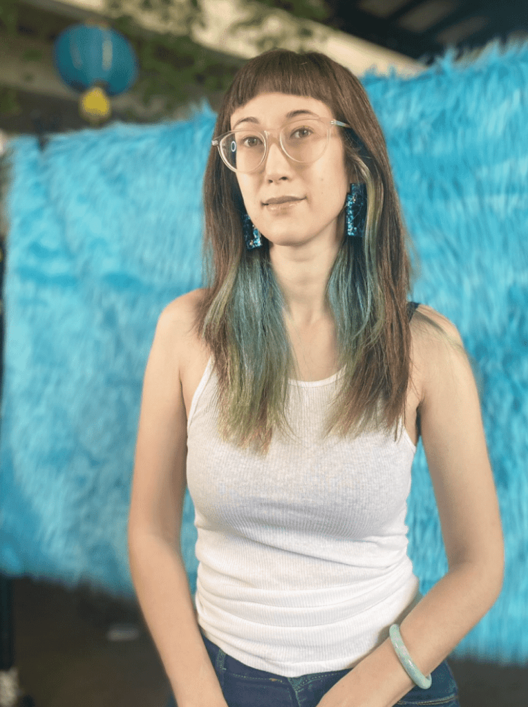 Meghan Weinstein stands in front of a furry blue background. She wears a white tank top and large blue earrings.