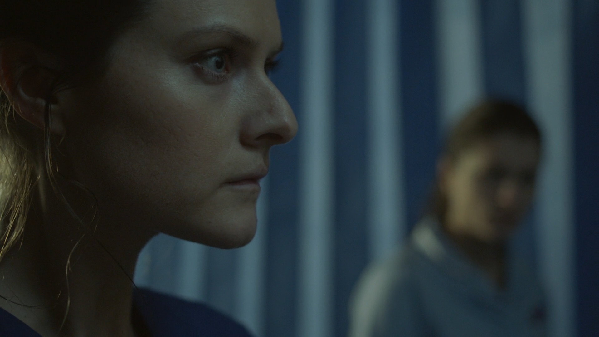 A young woman wearing nurse scrubs seen in profile view. Another woman is out of focus, standing in front of curtains.