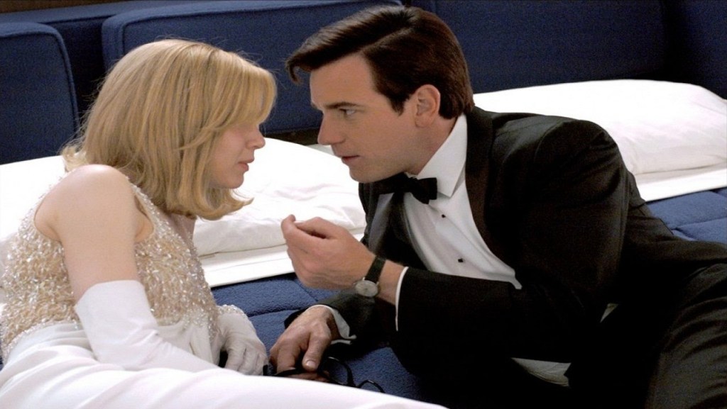 A woman and a man laying on a bed. The woman wears a fancy white dress and gloves and has strawberry blonde hair. The man wears a black tuxedo, seeming to be in the middle of an intense conversation with her.