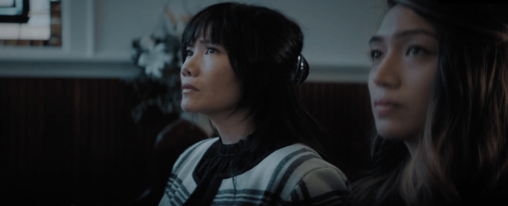 Two young Asian girls sit at a funeral while looking at something. The girl on the right is out of focus, and the girl on the left has short dark hair and a white striped top. White flowers can be seen in the background.