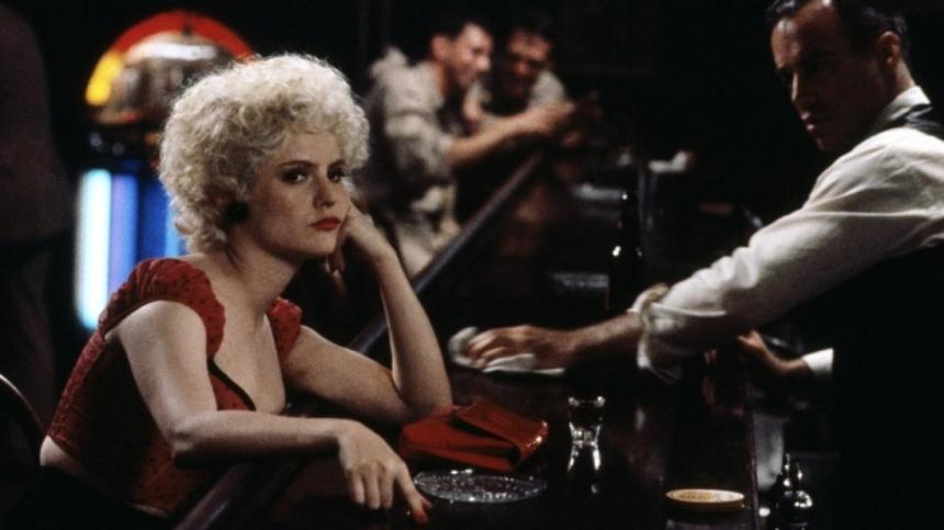 Jennifer Jason Leigh sits at a bar with her head resting on her hand. A bartender looks at her as he wipes down the counter.