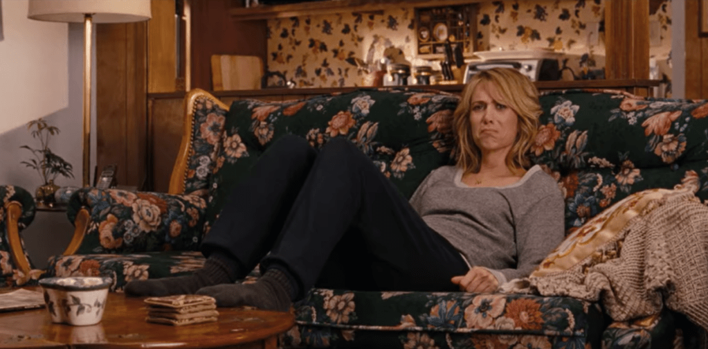 A white woman with blonde hair sits on a floral couch. She has an upset expression and wears casual clothes, with her legs up in front of her. A floral wallpaper can be seen behind her, and her feet rest on a wooden table where a floral cup sits.