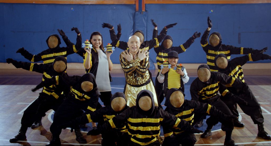A middle-aged bald man stands in the center of a room, smiling with his arms crossed. He is surrounded by dancers, with faces hidden as they wear dark blue hoodies with yellow stripes. Two young people stand by him, holding up objects that look like bees.
