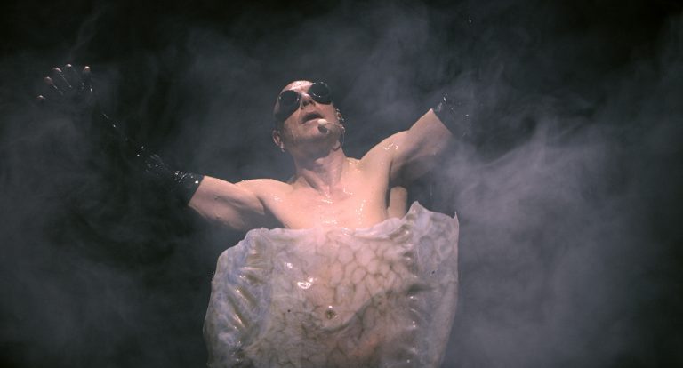 A middle-aged man with black over his eyes and black gloves. The lower half of his body is wrapped in something, and the background is obscured by smoke.
