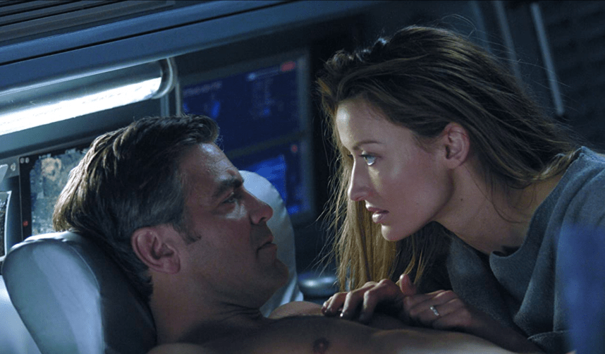 Natasha McElhone leans close to George Clooney as he lays in a reclining chair.