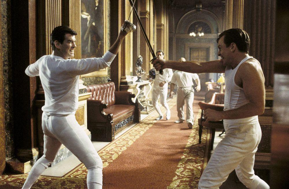 Pierce Brosnan as James Bond fences with Toby Stephens as Gustav Graves in an ornate, palace-like hallway. Two men, also in fencing outfits, come running toward them.